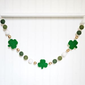 May include: A St. Patrick's Day garland featuring green felt shamrocks, white and green felt balls, and gold beads. The garland is hanging against a white background.