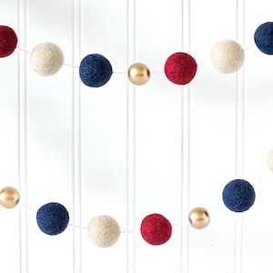 May include: A decorative garland featuring red, navy blue, cream, and gold-colored felt balls. The garland is strung on a thin white string, creating a festive and colorful display. The felt balls are evenly spaced.