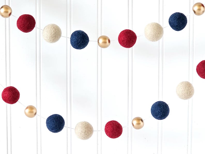May include: A decorative garland featuring red, navy blue, cream, and gold-colored felt balls. The garland is strung on a thin white string, creating a festive and colorful display. The felt balls are evenly spaced.