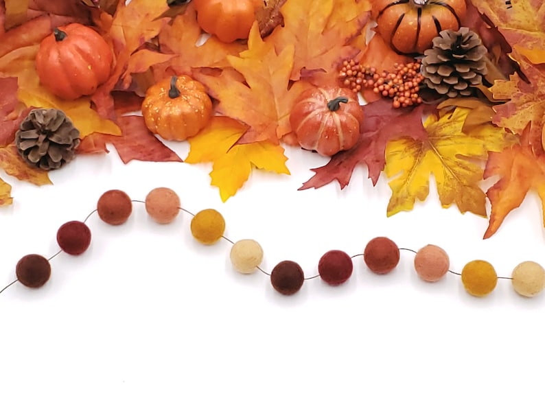 May include: A garland of felt balls in shades of orange, brown, and cream. The garland is hanging against a white background. The garland is decorated with fall leaves and pumpkins.
