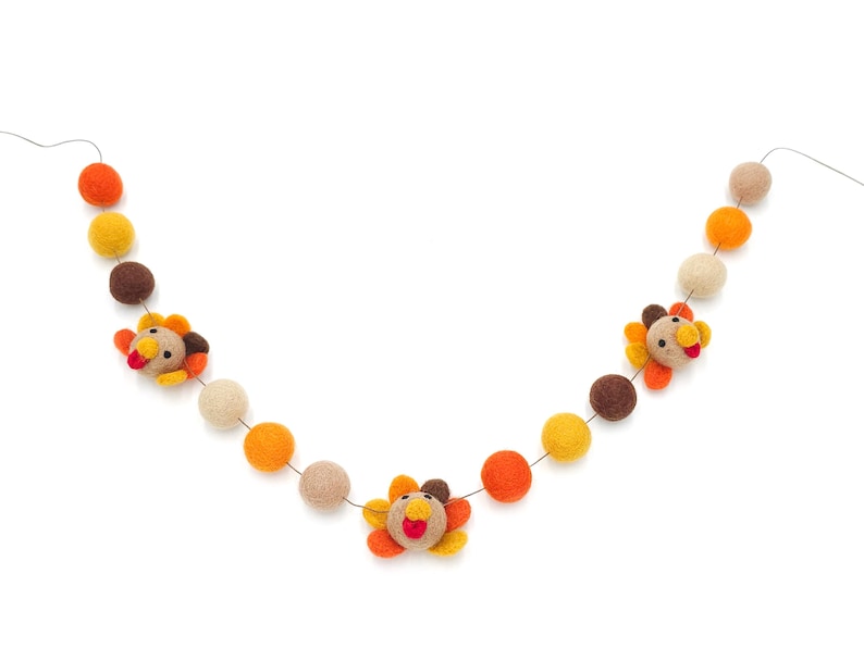 May include: A garland with felt balls in orange, brown, and beige colors. The garland features three felt turkey decorations with orange, brown, and yellow accents.