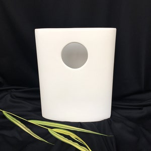 May include: A white, cylindrical container with a circular cutout. The container is set against a black backdrop, with a sprig of green and yellow leaves in the foreground. The object has a minimalist design.