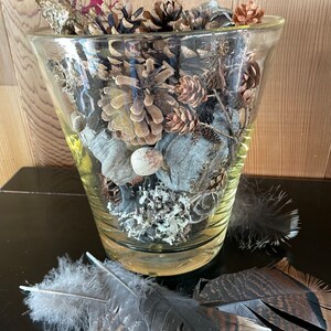 Op de afbeelding: Een glazen vaas gevuld met dennenappels, gedroogde bloemen en veren. De vaas staat op een zwart oppervlak.