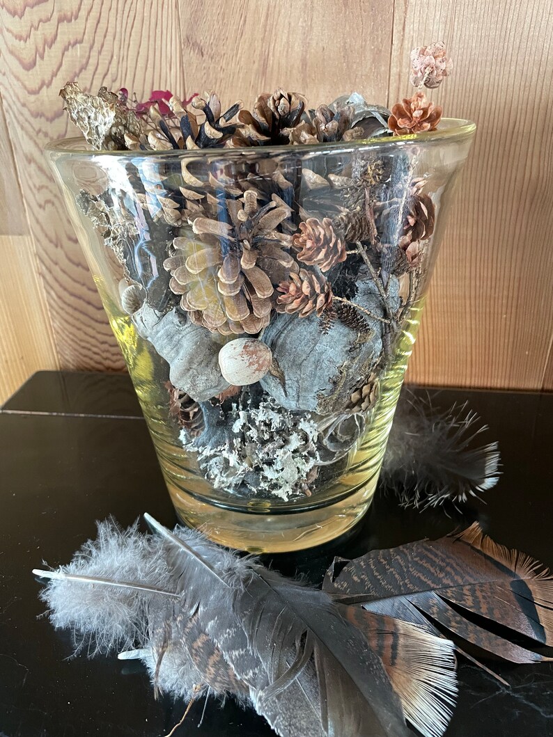Op de afbeelding: Een glazen vaas gevuld met dennenappels, gedroogde bloemen en veren. De vaas staat op een zwart oppervlak.