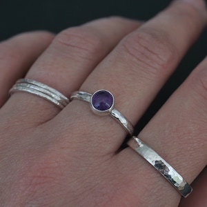 Puede incluir: Una mano con tres anillos de plata, uno con una piedra preciosa púrpura. Los anillos son texturizados y tienen un acabado martillado.