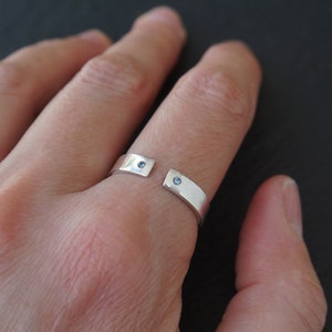 Peut inclure: Une bague en argent avec deux petites pierres bleues serties dans la bande. La bague est ouverte d'un côté et est portée sur un doigt.