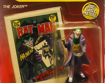 Könnte beinhalten: Eine Druckgussfigur des Jokers aus DC Comics. Der Joker trägt einen lila Anzug mit einem grünen Hemd und einer grünen Krawatte. Er hält eine Spielkarte in seiner rechten Hand und eine Lupe in seiner linken Hand. Die Figur steht auf einem schwarzen Sockel. Die Verpackung enthält ein Comic-Cover mit dem Joker und Batman.