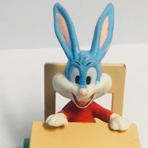 Könnte beinhalten: Eine blaue und weiße Comic-Kaninchenfigur, die an einem kleinen Holztisch mit brauner Platte und grünen Rädern sitzt. Das Kaninchen trägt ein rotes Hemd und hat ein breites Lächeln.