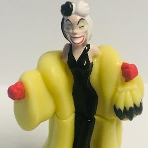 Peut inclure: Une figurine en plastique jaune de Cruella d'Enfer de 101 Dalmatiens de Disney. Elle porte une robe noire et un manteau de fourrure blanc et noir. Elle a les cheveux noirs et blancs et des gants rouges.