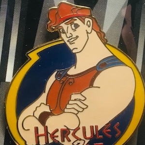 Könnte beinhalten: Eine gelbe, blaue und rote Emaille-Anstecknadel mit einem Cartoon-Bild von Herkules aus dem Disney-Film Herkules. Die Anstecknadel trägt den Text "Hercules 1997".