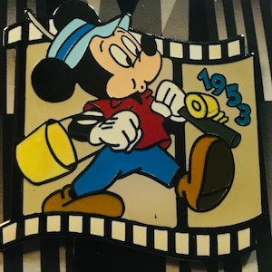 Könnte beinhalten: Eine Zeichentrick-Anstecknadel mit Mickey Mouse, der als Filmregisseur verkleidet ist und eine Filmklappe mit dem Jahr 1953 in der Hand hält. Die Anstecknadel ist vor einem Filmstreifen-Hintergrund platziert.