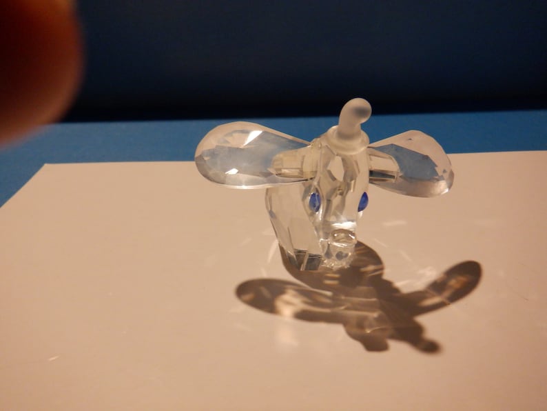 Puede incluir: Una figurita de elefante de cristal transparente con ojos azules. El elefante est&aacute; de pie sobre una superficie blanca.