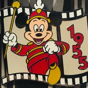 Może przedstawiać: Pin z czerwono-żółtym Mickey Mouse'em z obramowaniem w postaci taśmy filmowej. Mickey jest ubrany jak dyrygent i trzyma pałeczkę. Na pinie nadrukowany jest rok 1935.