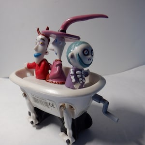 Pode incluir: Uma banheira de brinquedo branca de corda com três personagens do filme O Estranho Mundo de Jack. Os personagens são Jack Skellington, Sally e Oogie Boogie. O brinquedo tem uma manivela no lado para dar corda.