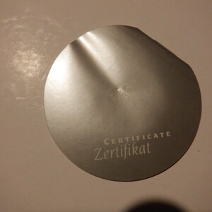 Puede incluir: Un certificado redondo plateado con el texto "CERTIFICATE Zertifikat" impreso en &eacute;l.