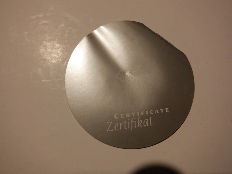 Puede incluir: Un certificado redondo plateado con el texto "CERTIFICATE Zertifikat" impreso en &eacute;l.