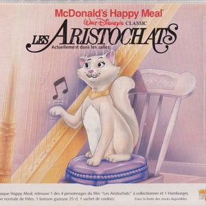 Puede incluir: Un gato blanco con un collar dorado toca un arpa. El gato está sentado en un cojín morado. La imagen es de una promoción de McDonald's Happy Meal para la película de Disney "Los Aristogatos".