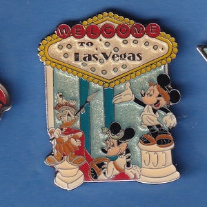 May include: A pin featuring Mickey Mouse, Donald Duck, and Goofy in front of a yellow and red "Welcome to Las Vegas" sign. The characters are wearing costumes and standing on pedestals.