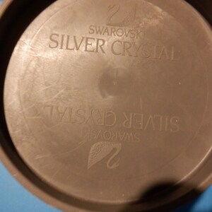 Puede incluir: Un recipiente redondo de pl&aacute;stico gris con el texto "Swarovski Silver Crystal" impreso en la parte superior e inferior.