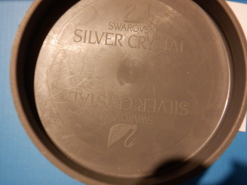 Puede incluir: Un recipiente redondo de pl&aacute;stico gris con el texto "Swarovski Silver Crystal" impreso en la parte superior e inferior.