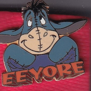 Könnte beinhalten: Eine blaue Eeyore-Anstecknadel mit einem gestickten Gesicht und dem Wort "Eeyore" in orangefarbenen Buchstaben. Die Anstecknadel befindet sich auf einem roten Hintergrund.