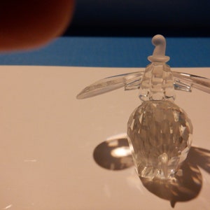 Puede incluir: Una figurita de ping&uuml;ino de cristal transparente con un sombrero blanco. El ping&uuml;ino est&aacute; de pie sobre una superficie blanca.