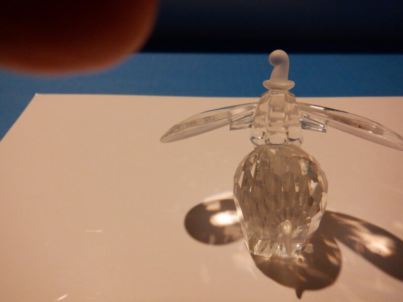 Puede incluir: Una figurita de ping&uuml;ino de cristal transparente con un sombrero blanco. El ping&uuml;ino est&aacute; de pie sobre una superficie blanca.