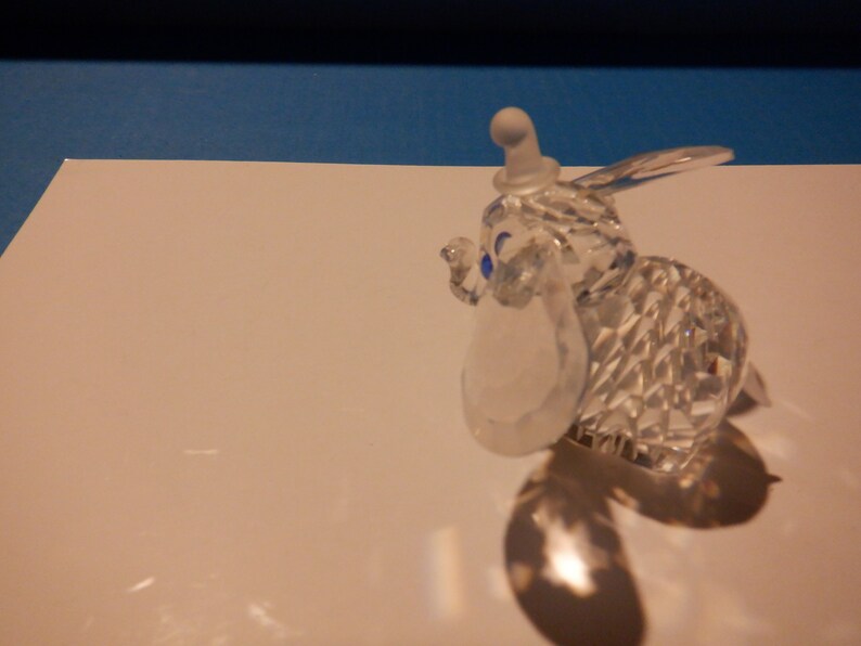 Puede incluir: Una figurita de cristal de Dumbo el elefante de la pel&iacute;cula animada de Disney. La figurita es transparente con ojos azules y un sombrero blanco.