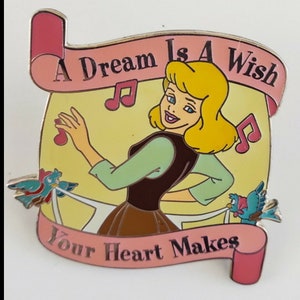 May include: A pink and yellow enamel pin featuring a cartoon image of Cinderella singing with a musical note in her hand. The text on the pin reads "A Dream Is A Wish Your Heart Makes".