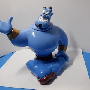 Puede incluir: Una figura de genio de cerámica azul de la película de Disney Aladdin. El genio está sentado con el brazo derecho extendido y la mano izquierda apoyada en la rodilla. Lleva un brazalete dorado en la muñeca derecha y una faja marrón alrededor de la cintura.