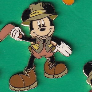 May include: A cartoon Mickey Mouse wearing a brown explorer hat and a green vest with a brown and white patterned design. He is holding a brown stick in his left hand and has a white glove on his right hand.