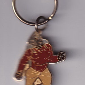 May include: A gold-colored metal keychain featuring a cartoon figure of a person wearing a red jacket and tan pants. The figure is running and has a determined expression on its face.