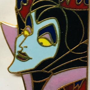 Op de afbeelding: Een close-up van een zwarte en gouden emaille pin met een cartoon afbeelding van Maleficent uit Disneys Doornroosje. De pin heeft een rood, geel en blauw kleurenschema en toont Maleficents kenmerkende hoorns en spitse kin.