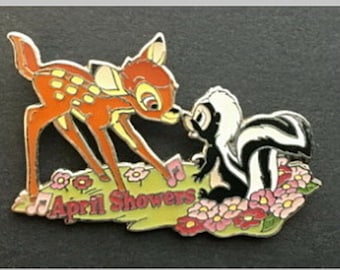 Peut inclure: Une épingle représentant Bambi le cerf et Fleur la moufette du film de Disney Bambi. Bambi est orange et brun avec des taches blanches. Fleur est noir et blanc avec une longue queue. Ils se tiennent sur un patch d'herbe verte avec des fleurs roses et le texte "April Showers".