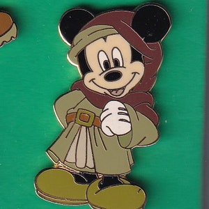May include: A cartoon pin of Mickey Mouse dressed as a medieval peasant. He is wearing a brown tunic with a tan sash and a brown hood. He is smiling and holding his hands together in front of him.