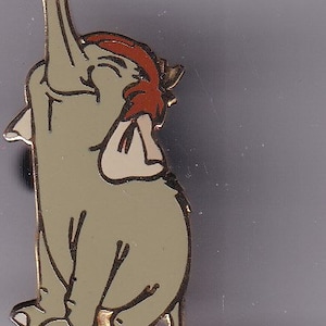 May include: A cartoon-style elephant pin with a light green body, red head covering, and white accents. The elephant is sitting with its trunk raised. The pin has a gold-colored border.