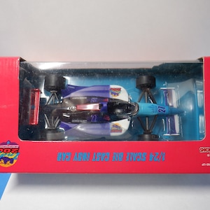 Puede incluir: Un coche Indy de fundición a escala 1/24 en un esquema de colores rojo, blanco y azul con el número 27 en el lateral. El coche está en una vitrina de plástico transparente sobre una caja roja con las palabras "Indy 500" y "Champions Race Cars" impresas en ella.