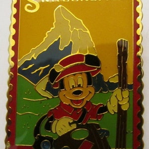 May include: A gold-colored pin featuring Mickey Mouse wearing a red hat and a red shirt. He is holding a walking stick and a brown bag with a red diamond on it. The background is a yellow and green landscape with a mountain and the word "SWITZERLAND" at the top.