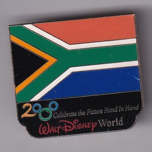 May include: A black and white pin with the South African flag and the text "2000 Celebrate the Future Hand in Hand Walt Disney World".