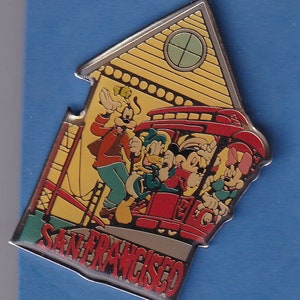 May include: A colorful enamel pin featuring Mickey Mouse, Donald Duck, Goofy, and Minnie Mouse riding a red cable car in San Francisco. The pin is shaped like a house with a yellow facade and a window. The text "San Francisco" is written in red and white letters at the bottom of the pin.