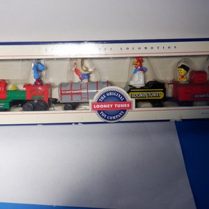 May include: A vintage Looney Tunes toy train set in its original box. The train features a green and red engine, a grey boxcar, a black flatcar, and a red caboose. Each car features a different Looney Tunes character: Bugs Bunny, Daffy Duck, Sylvester the Cat, and Tweety Bird. The box is white with blue trim and features the Looney Tunes logo.