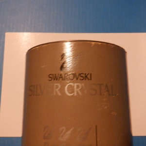 Puede incluir: Una caja cil&iacute;ndrica marr&oacute;n con el texto "SWAROVSKI SILVER CRYSTAL" impreso en la parte delantera. La caja tiene un logotipo de cisne plateado.