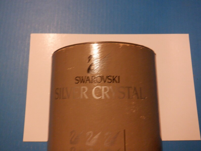 Puede incluir: Una caja cil&iacute;ndrica marr&oacute;n con el texto "SWAROVSKI SILVER CRYSTAL" impreso en la parte delantera. La caja tiene un logotipo de cisne plateado.