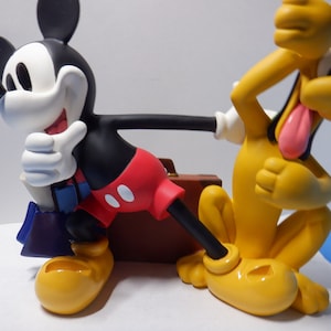 Peut inclure: Un chien de dessin animé jaune et noir avec une langue rouge qui sort tient une valise brune. Une souris de dessin animé noire et blanche avec un short rouge tient la valise et fait un pouce en l'air.