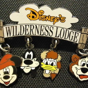 Puede incluir: Un pin de Disney's Wilderness Lodge con Mickey Mouse, Goofy, Donald Duck y un oso. El pin es blanco con texto negro y tiene forma de nube en la parte superior.
