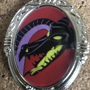 Op de afbeelding: Een zilveren ovaalvormige broche met een rood, paars en groen drakenontwerp. De draak is afgebeeld met een dreigende uitdrukking en scherpe tanden. De broche heeft een decoratief zilveren frame.