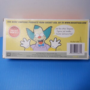 May include: A yellow box featuring a cartoon image of Krusty the Clown from The Simpsons. The box reads "Collect them all!" and "Another fine toy from Rocket USA".