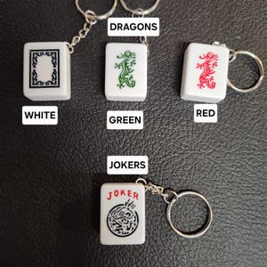 Puede incluir: Cuatro fichas de mahjong de cer&aacute;mica blanca con llaveros. Las fichas presentan un dise&ntilde;o de drag&oacute;n en verde, rojo y negro. Las fichas est&aacute;n etiquetadas como "DRAGON" y "JOKER".