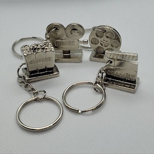 May include: A set of four silver keychains featuring movie-themed designs. The keychains include a film reel, a movie camera, a popcorn bucket, and a film strip. Each keychain has the text "Scream It 2" printed on it.