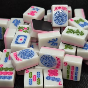 Puede incluir: Un conjunto de fichas de Mahjong blancas con bordes rosas. Las fichas presentan varios símbolos, números y la palabra "JOKER" en rosa. También hay diseños azules y verdes, creando un surtido colorido para el juego clásico.